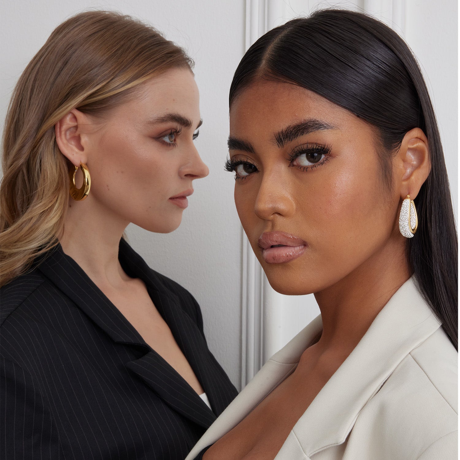 2 models wearing chunky hoop earrings. One wearing hoop earrings plated with 18K gold. The other wearing hoop earrings plated with rhodium and set with crystals.